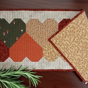 May include: A quilted table runner featuring four heart shapes in shades of green, orange, and tan. The hearts are arranged in a row against a light background. A matching placemat with a leaf pattern is folded to the side.
