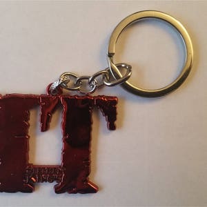 Peut inclure: Porte-clés en métal rouge avec les lettres "IT" dans une police de caractères stylisée. Le porte-clés est attaché à un anneau de clé argenté.
