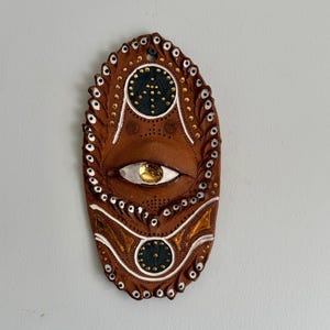 May include: Oval brown wall art with an eye design. The artwork features a central eye with a gold iris, surrounded by white and gold accents. The border is adorned with small white and gold dots, creating a decorative piece.