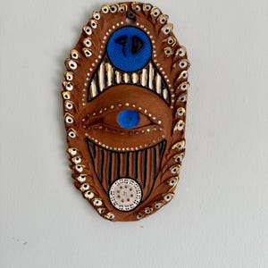 May include: A brown ceramic wall hanging with an eye design. The piece features a large eye with a blue iris, surrounded by smaller eye-shaped details with gold accents. The design includes white and black vertical lines and a blue circle.