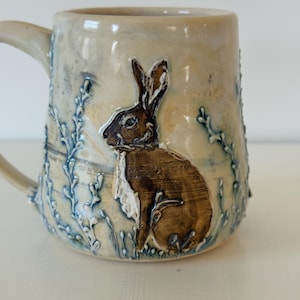 May include: A ceramic mug with a cream-coloured glaze featuring a raised, brown-toned relief of a rabbit sitting among blue-tinted, textured plant stalks. The mug has a curved handle on the left side and a smooth, rounded rim at the top.