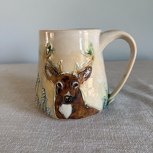 May include: Cream-colored ceramic mug featuring a detailed deer design. The deer is depicted with brown fur, antlers, and a white chest. The mug has a curved handle and is decorated with green and blue accents.