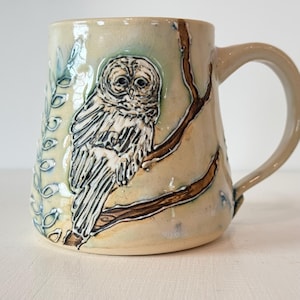May include: A ceramic mug with a cream-colored glaze featuring a raised, hand-carved relief of an owl perched on a brown branch. The design includes a vertical fern-like pattern on the left side, with subtle blue and green accents.