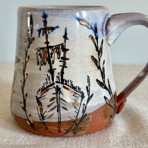 May include: A ceramic mug with a light gray upper section and a reddish-brown base. The mug features a hand-painted black and metallic gold illustration of a sailing ship centered on the front, flanked by vertical leafy branch designs.