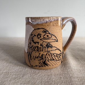 May include: A tan ceramic mug featuring a black raven illustration. The mug has a curved handle and a white and brown rim. The raven is drawn in a simple, artistic style.