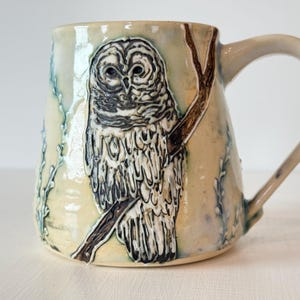 May include: A ceramic mug with a cream-colored glaze featuring a raised, textured relief of a barred owl perched on a brown branch. The owl has detailed black and white plumage, and the mug is decorated with subtle blue-green sprigs.