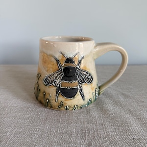 May include: Cream-colored ceramic mug featuring a detailed illustration of a bumblebee. The bee is black and yellow, with intricate wing details. The mug has a curved handle and is decorated with small floral accents.