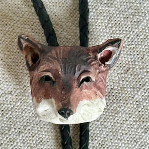 May include: A bolo tie featuring a sculpted fox head pendant with brown, black, and white painted details. The fox head is attached to a black braided leather cord that extends upwards and downwards from the center of the animal figure.