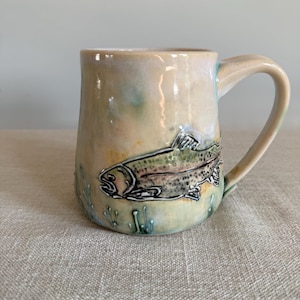 Pode incluir: Uma caneca de cerâmica com um esmalte brilhante em tons de bege e verde pálido. O exterior apresenta uma ilustração detalhada desenhada à mão de uma truta a nadar entre plantas aquáticas estilizadas, feita com tinta preta e subtis toques de cor.