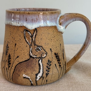 May include: A handmade ceramic mug with a brown speckled finish. The mug features a detailed illustration of a rabbit and wheat stalks. The rim is a light purple color with a white glaze. The handle is curved and brown.