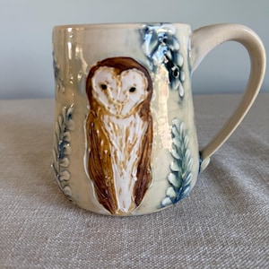 May include: Ceramic mug in cream with a detailed owl illustration. The owl is brown and white, with a textured finish. The mug has a curved handle and is decorated with blue and green leaf accents. A unique piece for the kitchen.