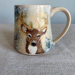 May include: A ceramic mug featuring a hand-painted brown deer with antlers centered on a cream-colored background. The design includes raised, textured leaf motifs in shades of blue and grey on the sides, with a curved handle on the right side.