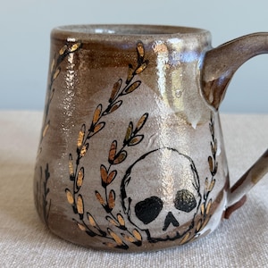 Könnte beinhalten: Ein Keramikbecher mit einer glänzenden, gesprenkelten beigen und braunen Glasur. Die Außenseite zeigt eine handgemalte schwarze Totenkopf-Illustration, umgeben von zarten, vertikalen Blattzweigen mit metallischen Goldblatt-Akzenten und einem Henkel.