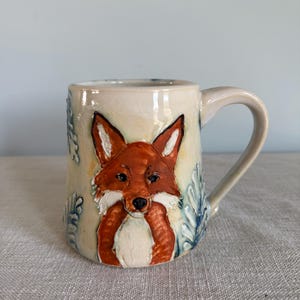 May include: A ceramic mug featuring a hand-painted fox. The fox is painted with orange and white fur, and the mug has blue leaf accents. The mug has a cream-coloured background and a curved handle.