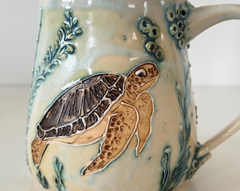 May include: A ceramic mug with a light blue and cream glaze featuring a raised, textured relief of a sea turtle swimming among seaweed and bubbles. The turtle has a dark brown shell and tan skin, with intricate carved details on the surface.