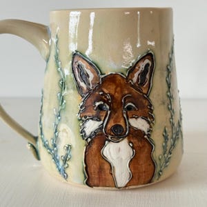 May include: A ceramic mug with a glossy cream-colored glaze featuring a raised, hand-painted fox illustration in shades of brown, white, and black. The design includes embossed vine-like patterns in a muted blue-green hue on the sides.