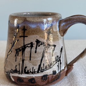 May include: A ceramic mug with a tapered shape and a curved handle. The exterior features a speckled tan and brown glaze with a blue-tinted rim. A black ink-style illustration of a sailing ship with masts and rigging is painted on the side.