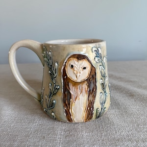 May include: A ceramic mug with a cream-colored glaze featuring a raised, textured relief of a brown and white barn owl centered on the front. The owl is flanked by embossed, leafy vine patterns in a muted green tone, and the mug has a curved handle.