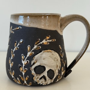 Może przedstawiać: Ceramiczny kubek z matowym czarnym wykończeniem, ozdobiony ręcznie malowaną białą czaszką i botanicznymi gałązkami ze złotymi akcentami. Górna krawędź i uchwyt są pokryte gładką, jasnobeżową glazurą, kontrastującą z ciemnym korpusem naczynia.