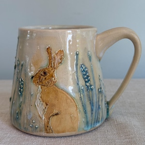 May include: A beige ceramic mug with a brown rabbit design and blue floral accents. The mug has a curved handle and a slightly textured surface. The rabbit is sitting in a field of blue flowers.
