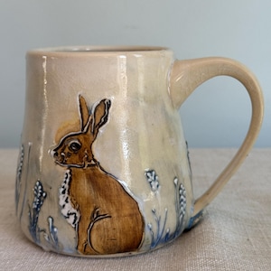 May include: A ceramic mug with a brown rabbit illustration. The mug has a beige background with blue floral accents. The handle is curved and the mug is approximately 4 inches tall.