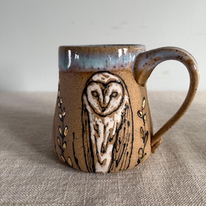 May include: A ceramic mug with a brown base and a light blue rim. The mug features a hand-painted owl design in black and white, with gold accents. The handle is curved and the mug is suitable for hot beverages.