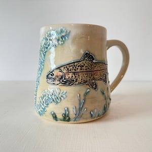 Op de afbeelding: Een beige keramieken mok met een verhoogd, getextureerd reliëf van een gespikkelde forel die tussen blauwgroene waterplanten zwemt. De mok heeft een glanzende afwerking, een gebogen handvat aan de rechterkant en een cilindervormig lichaam.