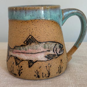 Puede incluir: Una taza de cerámica hecha a mano con un cuerpo moteado marrón y un borde y asa turquesa. La taza presenta una ilustración pintada a mano de un pez con detalles rosas y negros, y un pequeño diseño de planta en la parte inferior.