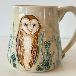 Puede incluir: Una taza de cerámica con esmalte beige brillante que presenta una lechuza en relieve pintada a mano en tonos marrones y blancos. El diseño incluye tallos florales y botánicos de color azul verdoso en relieve alrededor de la lechuza, con un asa.