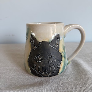May include: A ceramic mug featuring a detailed black wolf face. The mug has a beige glaze with blue and green accents. The handle is curved, and the mug appears to be handmade.