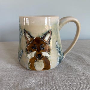 May include: A ceramic mug with a hand-painted fox design. The mug features a brown fox with white accents on a cream-colored background. The mug has a curved handle and is decorated with blue and green leaf patterns.