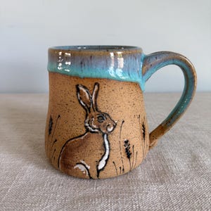 Może przedstawiać: Ręcznie robiony ceramiczny kubek z brązowym, nakrapianym wykończeniem. Kubek przedstawia szczegółową ilustrację zająca na polu pszenicy. Obręcz i uchwyt są glazurowane w odcieniach niebieskiego, dodając koloru do rustykalnego designu.
