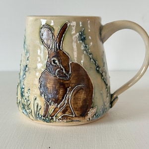 May include: A ceramic mug with a glossy cream-colored glaze featuring a hand-painted brown rabbit in the center. The design includes raised, textured blue-green botanical elements surrounding the rabbit, with a curved handle on the right side.