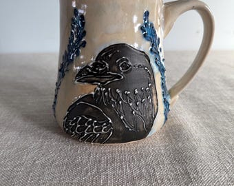May include: A ceramic mug with a bird design. The mug features a beige background with a black bird illustration on the front. Blue leaf-like accents are on either side of the bird. The handle is beige.