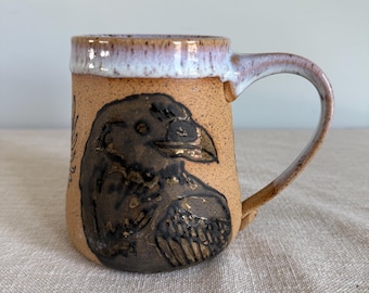 May include: A handmade ceramic mug with a raven design. The mug features a tan base color with a dark brown raven illustration and a white and brown rim. The handle is curved and the mug is suitable for hot or cold beverages.