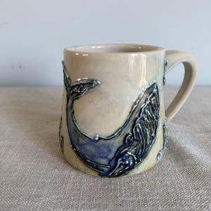 Pode incluir: Caneca de cerâmica creme com um design de baleia azul. A baleia é esculpida em relevo, com detalhes em azul mais escuro e prateado. A caneca tem uma alça curva e é adequada para bebidas quentes ou frias.