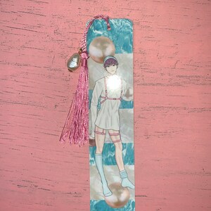 K&ouml;nnte beinhalten: Ein Lesezeichen mit einer Person mit rosa Haaren, die ein wei&szlig;es Hemd und wei&szlig;e Socken mit blau-wei&szlig;em Hintergrund tr&auml;gt. Das Lesezeichen hat einen rosa Quasten mit einem Perlenakzent.