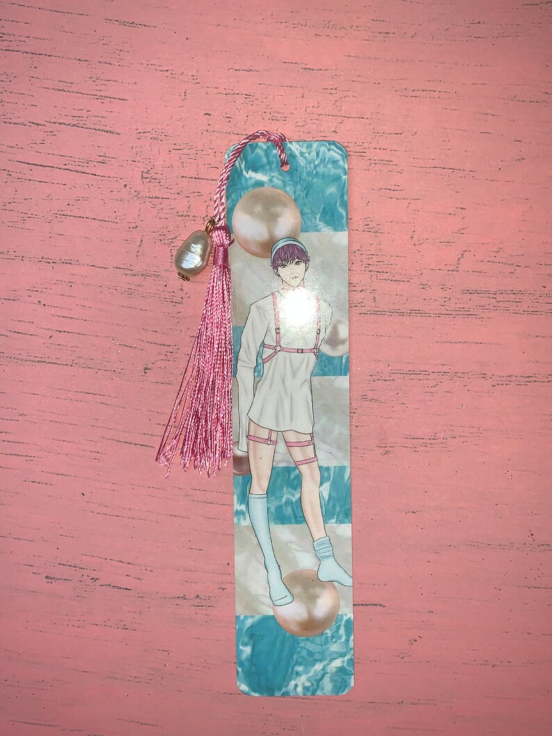 K&ouml;nnte beinhalten: Ein Lesezeichen mit einer Person mit rosa Haaren, die ein wei&szlig;es Hemd und wei&szlig;e Socken mit blau-wei&szlig;em Hintergrund tr&auml;gt. Das Lesezeichen hat einen rosa Quasten mit einem Perlenakzent.