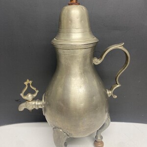 May include: Antique pewter tea or coffee pot with a wooden finial. The pot has a bulbous body, a curved handle, and a decorative spigot. It stands on three ornate legs with wooden feet. The lid is hinged and the pot has a classic, vintage design.