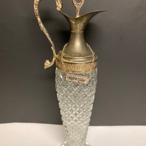 May include: A vintage glass pitcher with a silver-colored metal handle, spout, and lid. The clear glass body has a diamond pattern. A small tag with text is attached to the pitcher. The pitcher is approximately 12 inches tall.