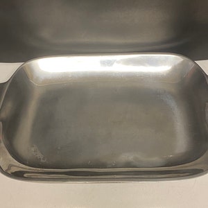 May include: A rectangular, silver-colored serving tray with ornate, scroll-like handles on each end. The tray has a slightly curved shape and a smooth surface, suitable for serving food or displaying items. The handles add a decorative touch to the tray's design.