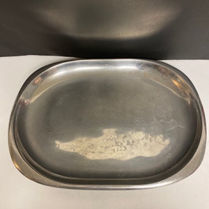 Könnte beinhalten: Ein ovales Serviertablett aus Edelstahl mit leicht erhöhtem Rand. Das Tablett hat eine gebürstete Metalloberfläche und weist einige Gebrauchsspuren auf. Die Form ist länglich, mit abgerundeten Enden und sanft geschwungenen Seiten.