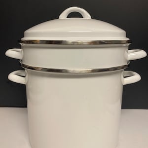 Könnte beinhalten: Ein weißer Emaille-Kochtopf mit Deckel und Dampfeinsatz. Der Topf hat zwei seitliche Griffe und einen silbernen Rand. Der Deckel hat einen Schlaufengriff. Der Topf ist zum Dämpfen oder Kochen von Speisen bestimmt.