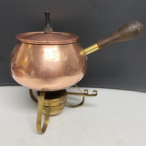 Peut inclure: Une marmite à fondue en cuivre avec un couvercle et une poignée en bois. Le pot repose sur un support en laiton avec des pieds incurvés. La poignée est en bois foncé. Le pot est rond et possède un petit bouton en bois foncé sur le couvercle.