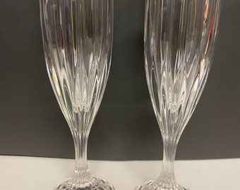 Può includere: Due flûte da champagne trasparenti con dettagli a coste verticali. Gli steli sono sottili e attorcigliati, che conducono a una base circolare con un motivo a raggi. Le flûte sono in vetro trasparente e sono su uno sfondo scuro.