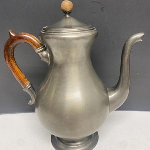 May include: A pewter teapot with a matte grey finish, featuring a curved wooden handle and a matching wooden finial on the lid. The vessel has a wide, rounded body, a pedestal base, and a long, slender spout extending from the right side.