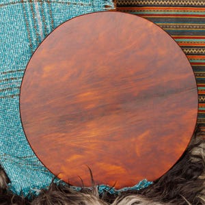May include: A large, round, wooden drum with a rich, reddish-brown finish. The drum's surface has a natural wood grain pattern. The drum is set against a backdrop of colorful textiles, including a turquoise woven fabric and a striped pattern.