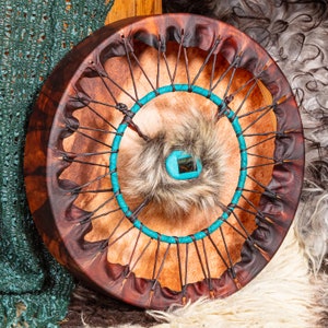 May include: A brown leather drum with a turquoise blue rope wrapped around the center. The drum has a fur patch in the center with a turquoise blue circle.