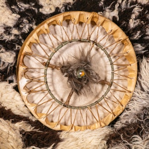 May include: A traditional Native American hand drum with a brown leather frame and a white hide stretched over it. The drum has a green and brown lacing pattern and a brown feather in the center.
