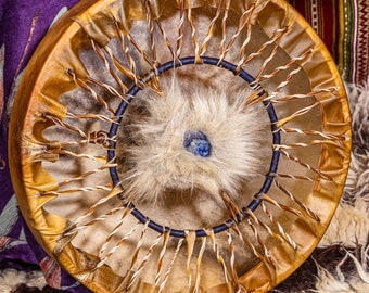 May include: A traditional Native American hand drum with a brown leather frame and a white fur center. The drum has a blue stone in the center of the fur.